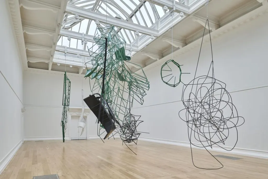 Twisted green and black metal sculptures hang beneath a gallery’s glass roof.