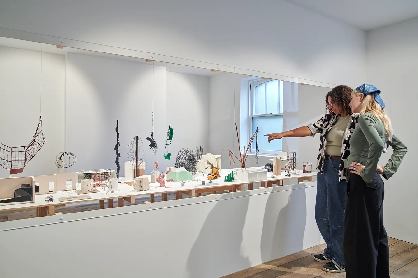 Two gallery visitors examining abstract sculptures behind glass, with one pointing towards a suspended piece.
